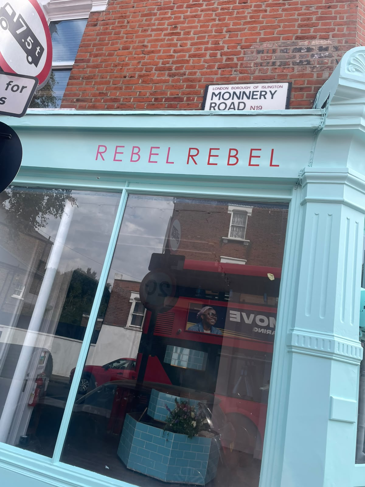 Rebel Rebel Pastel blue storefront with large glass window and pink'REBEL REBEL' sign, brick building behind. On Monnery Road, London.
