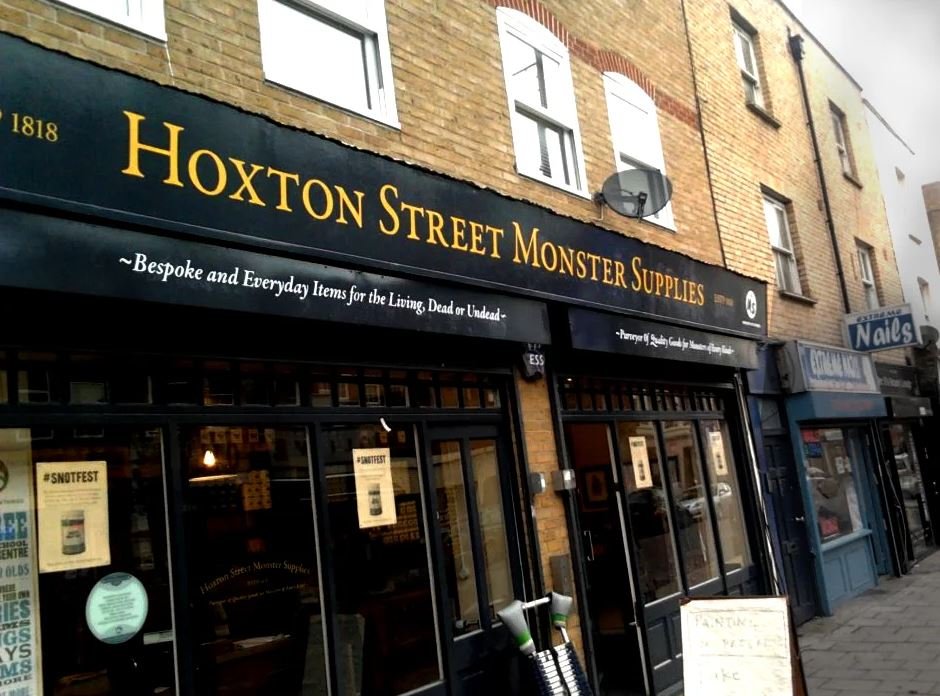 Storefront sign reading 'Hoxton Street Monster Supplies' on a brick building with a black awning and slogan underneath; windows and sidewalk in view.