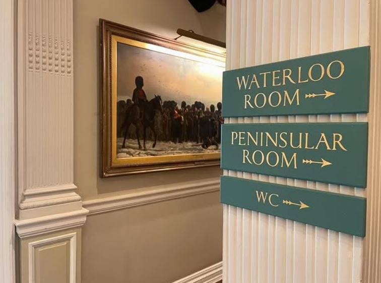 Interior hallway with teal wall signs reading 'WATERLOO ROOM', 'PENINSULAR ROOM', and 'WC' mounted on a white paneled column; a framed painting of riders and people on the wall to the left.