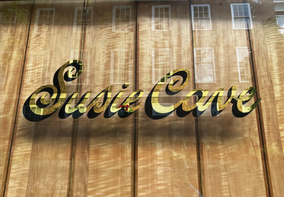 Sign reading'Susie Cares' in gold cursive lettering on a wood-paneled wall (with glass reflection).