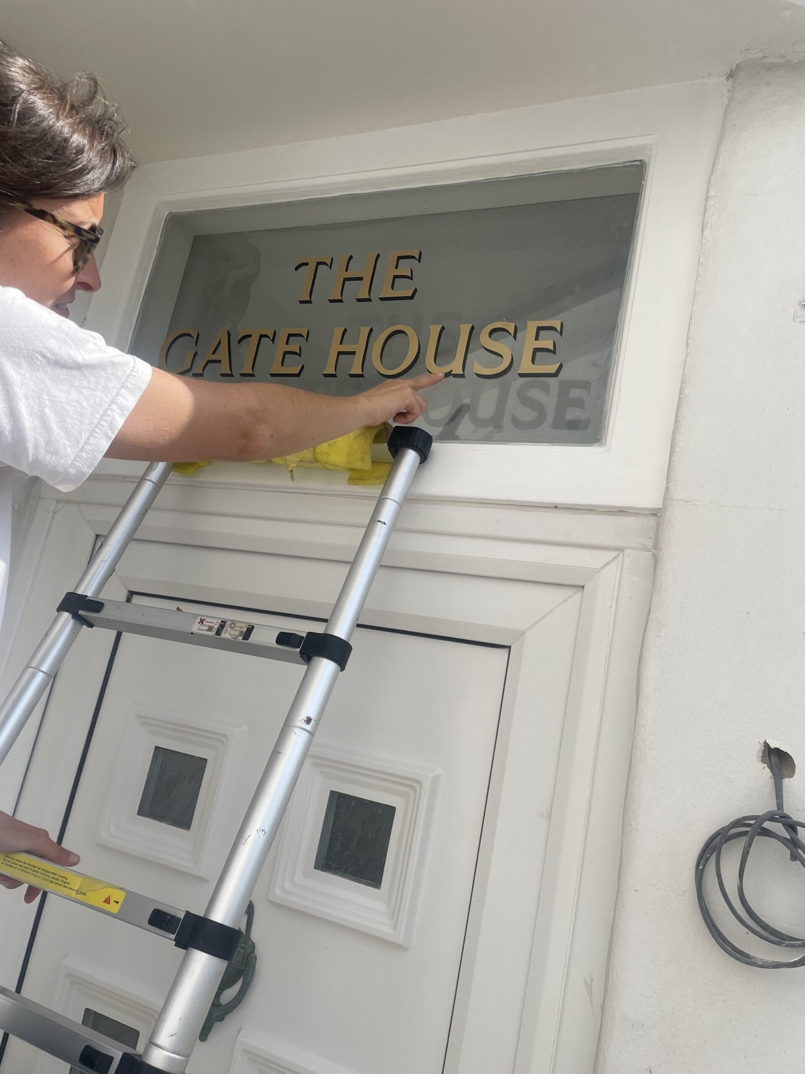 Sign Writing by Seraina Signwriters of London | NGS Sign lettering & writing. Person on a step ladder pointing at gold letters that spell'THE GATE HOUSE' on a glass window next to a white door frame.