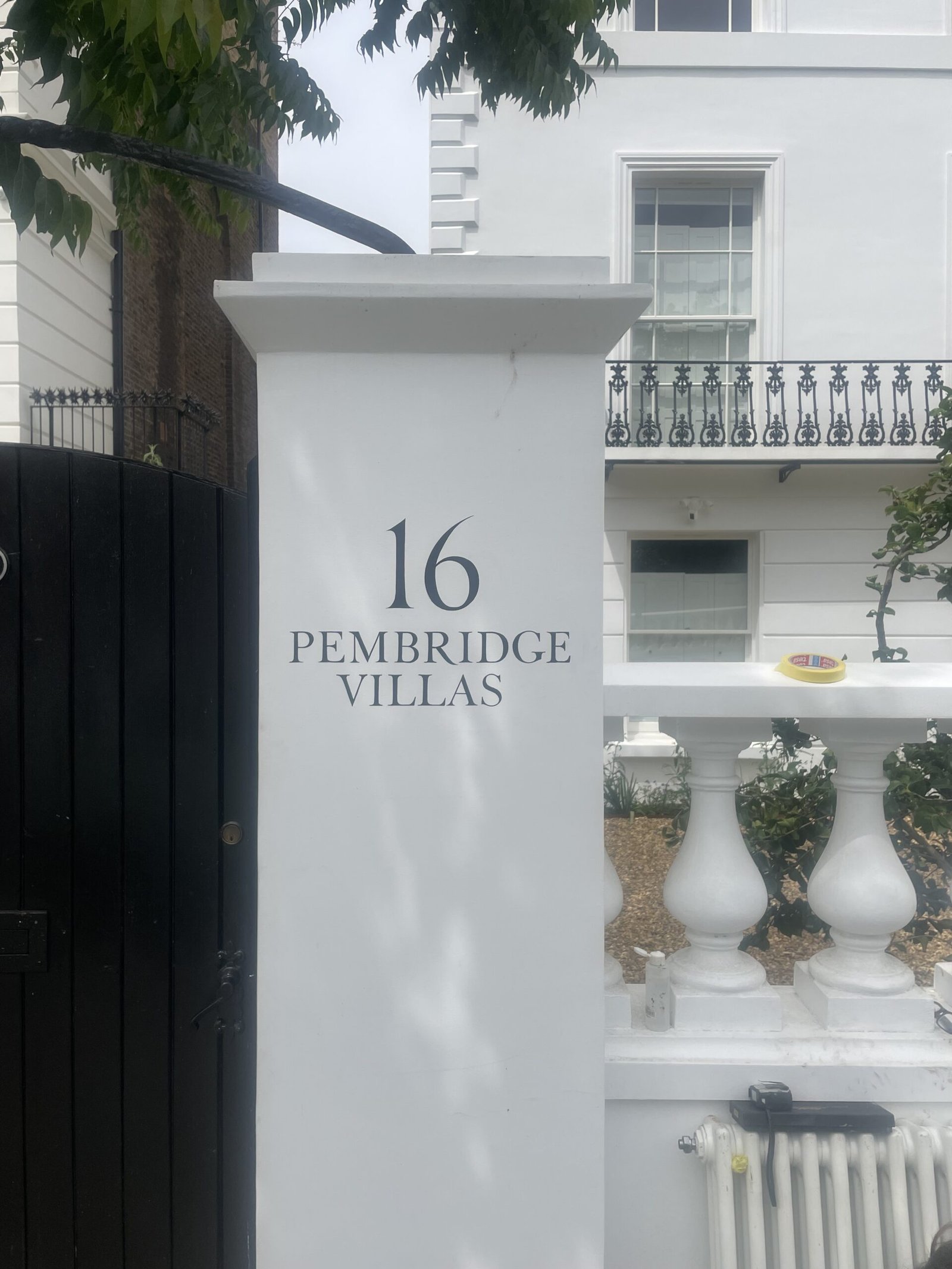 pillar number 16 Sign Writing by NGS Signwriters of London | NGS Sign lettering & writing. White pillar with the number 16 and'PEMBRIDGE VILLAS' sign at a gated townhouse entrance, with a decorative balcony and tree branches above.