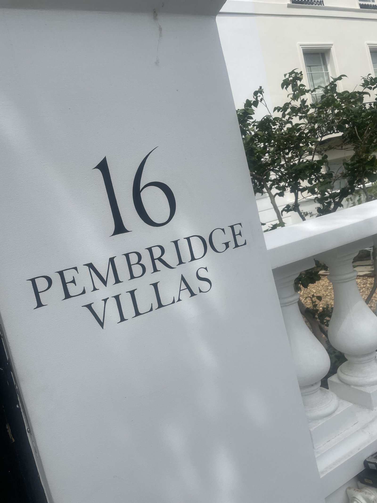 '16' 'PEMBRIDGE VILLAS'Sign Writing by Seraina Signwriters of London | NGS Sign lettering & writing. White outdoor sign with a large black number'16' and the words 'PEMBRIDGE VILLAS' visible, next to a white railing and green shrubbery.