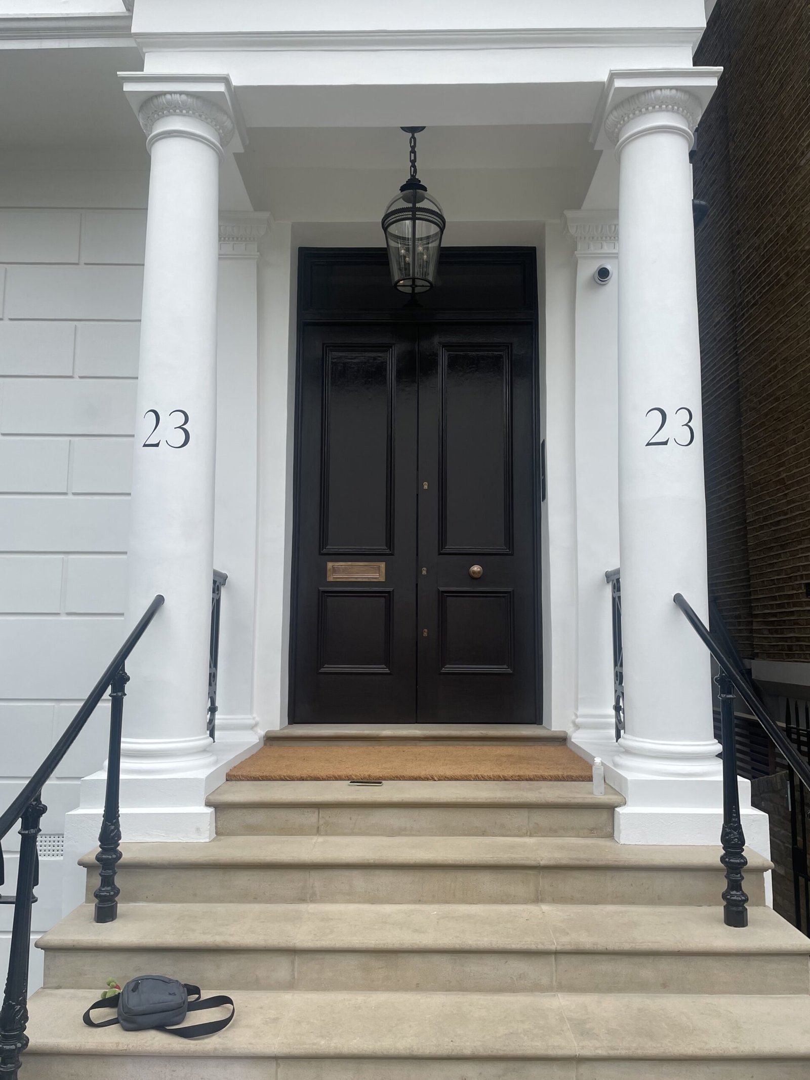 Sign Writing by NGS Signwriters of London | NGS Sign lettering & writing. White-columned townhouse entrance with a black double door, hanging lantern, and the number 23 on both columns; a backpack sits on the steps.