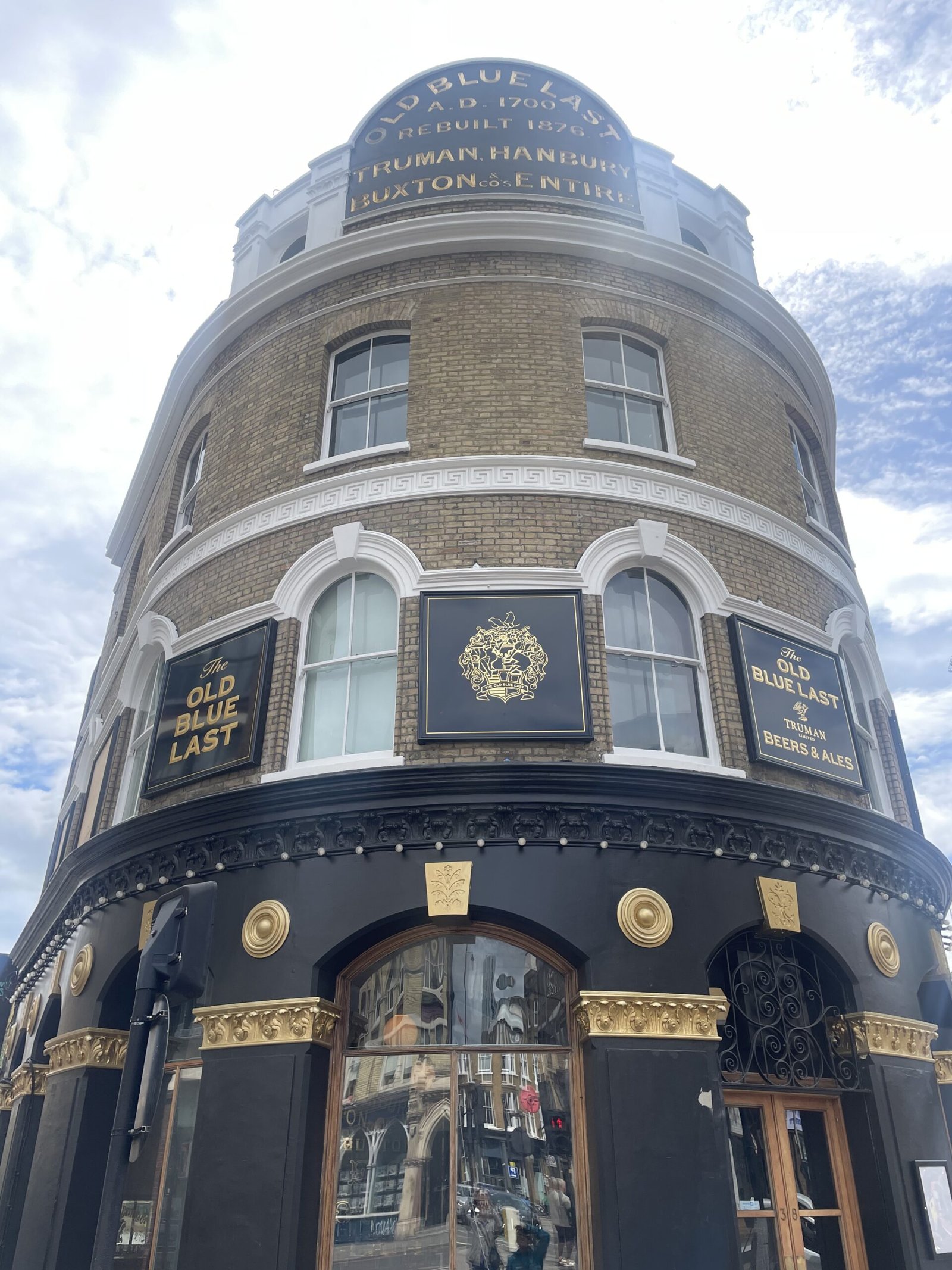 Corner view of The Old Blue Last pub, beige brick facade with white trim and black/gold signage reading 'The Old Blue Last' and decorative crest.'The Old Blue Last' Signage NGS and 'Since 1876' under an arched window.