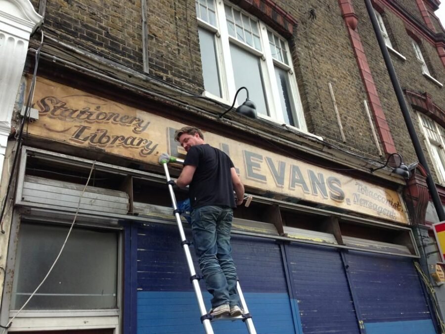 A man on a tall ladder repairs a faded shop sign above a storefront.