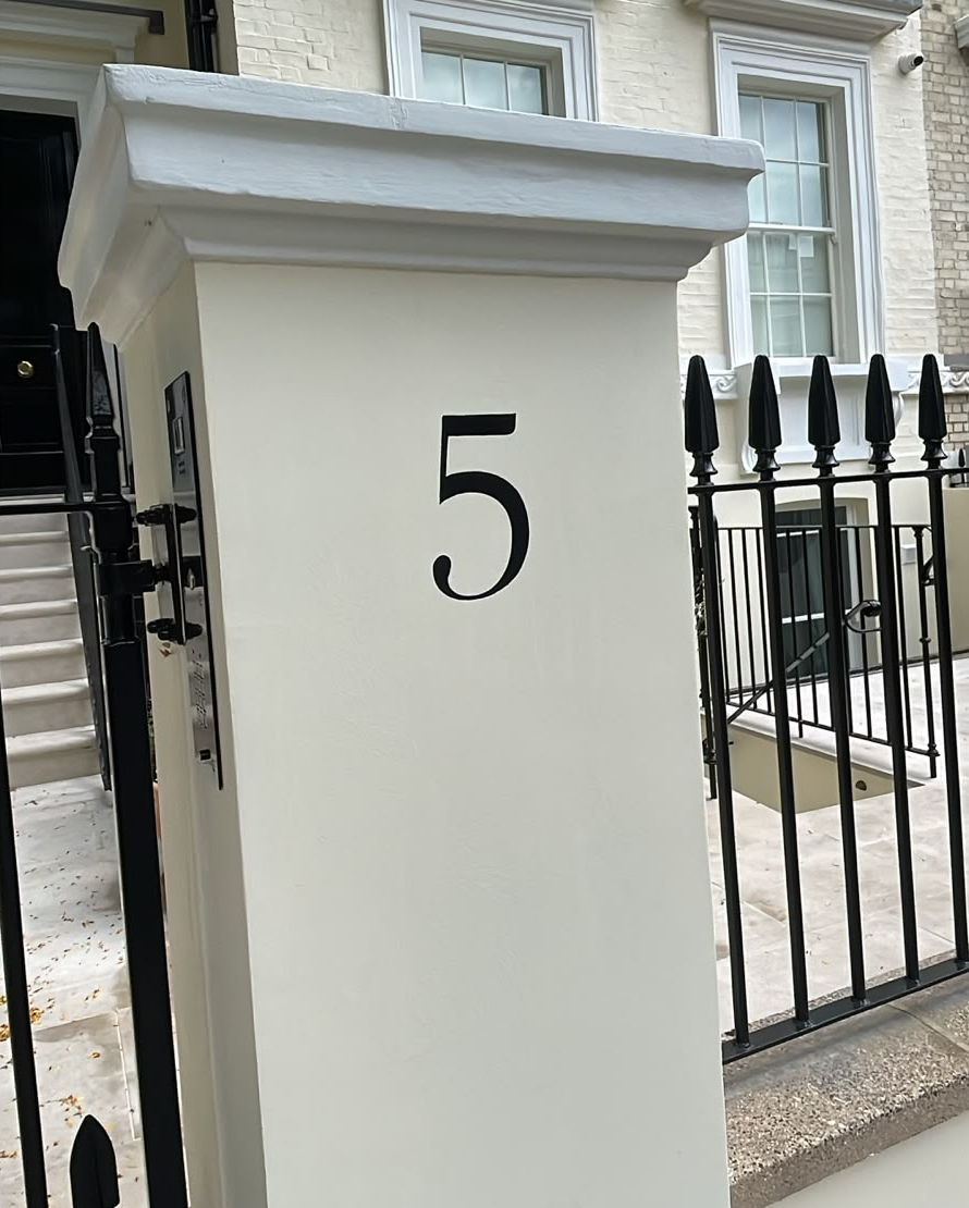 White front pillar with the house number 5 in black, next to a black wrought-iron gate.