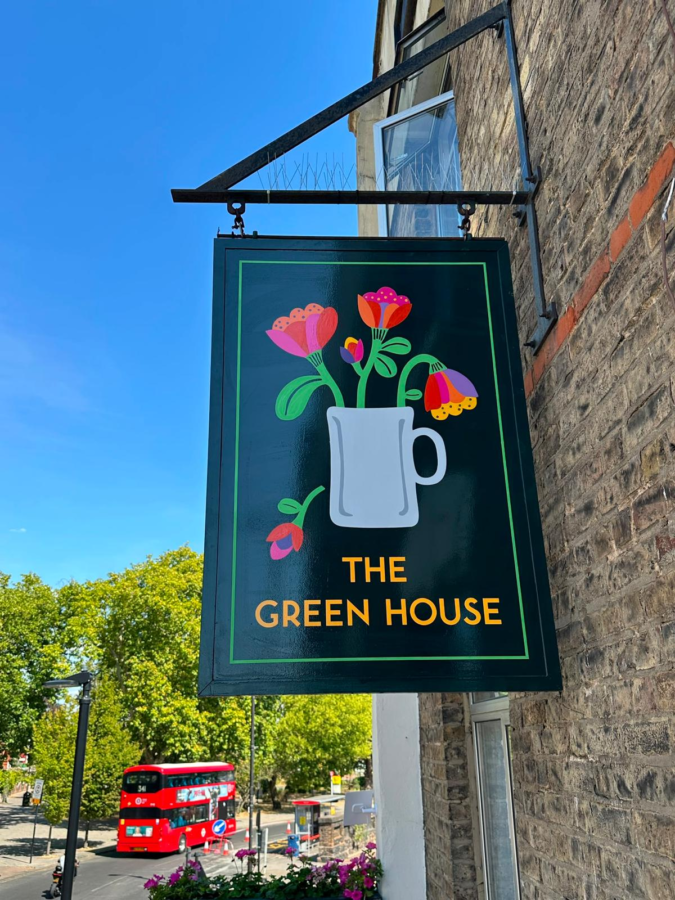 Signboard for 'The Green House' with a white pitcher and colorful flowers against a dark green background, hanging outside a brick building.